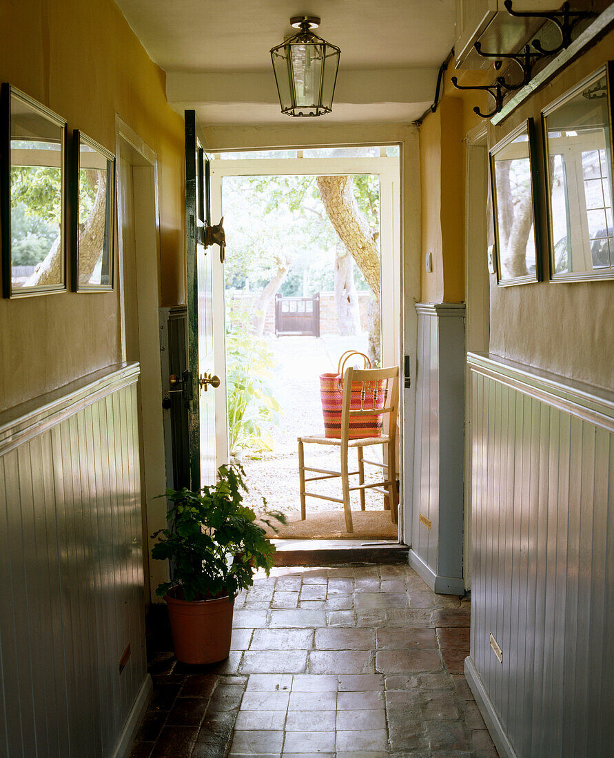Gelber Flur im Landhausstil mit gefliestem Boden, Holzvertäfelung, Lampe, Holzstuhl und geöffneter Vordertür zum Garten