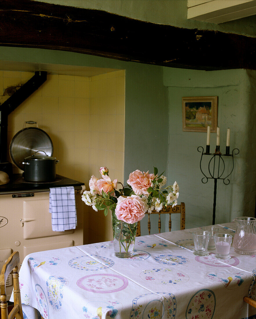 Gelbe Landhausküche mit Balkendecke, Esstisch mit Tischtuch und Blumenstrauß, dahinter Aga-Herd in einer Kachelnische