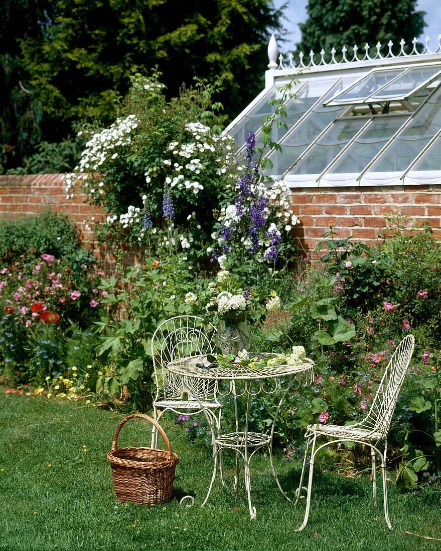 Traditioneller kleiner Metalltisch und Stühle in einem ummauerten englischen Landgarten mit Blumenrabatten und Gewächshaus