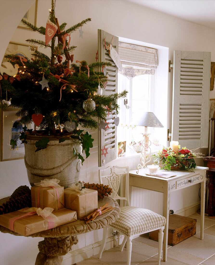 Detail eines geschmückten getopften Weihnachtsbaums auf einem Tisch, umgeben von Geschenken und einem Fenster mit Fensterläden über einem kleinen Beistelltisch mit einer Blumenauslage