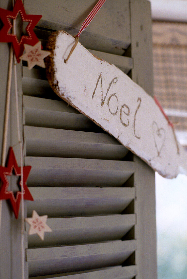 Handgeschnitztes Schild mit der Aufschrift 'Noel' aufgehängt an einem Fensterladen