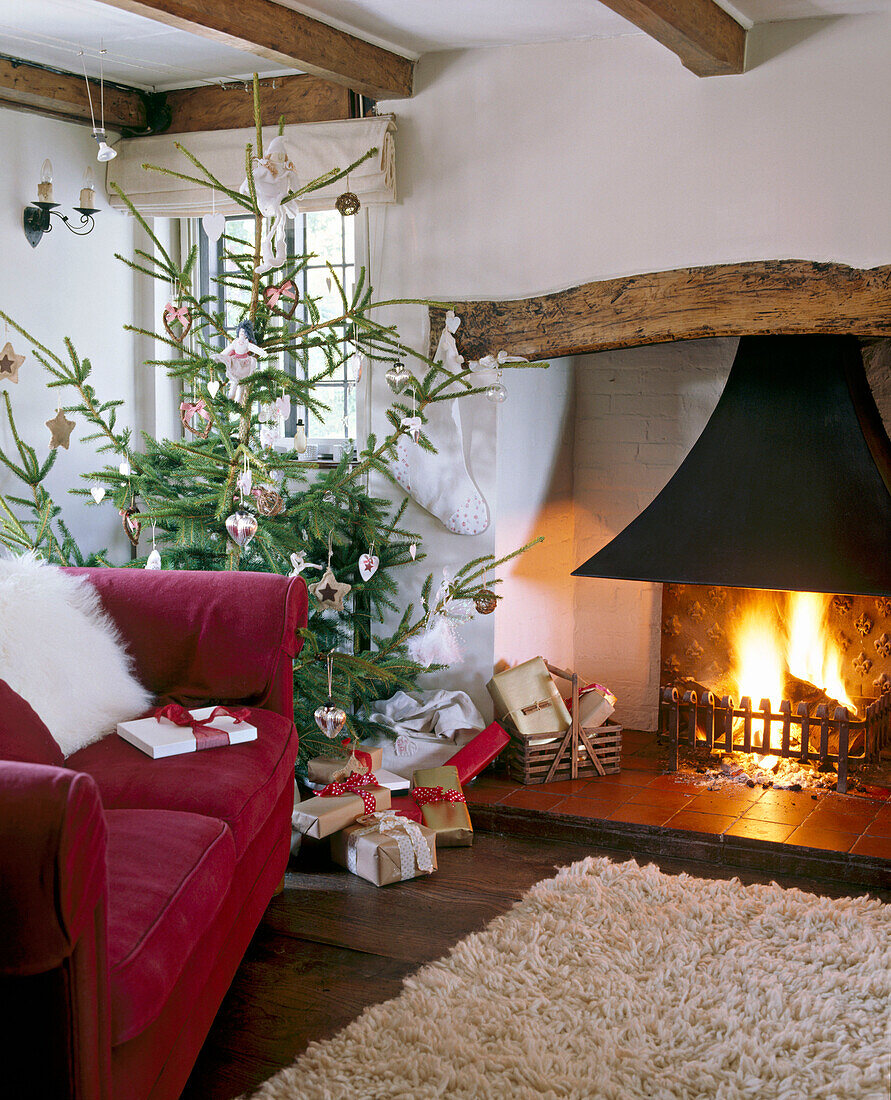 Gemütliches, weihnachtlich geschmücktes Wohnzimmer im Landhausstil mit Balkendecke, Kamin, geschmücktem Weihnachtsbaum mit Geschenken, Teppich und Polstersofa