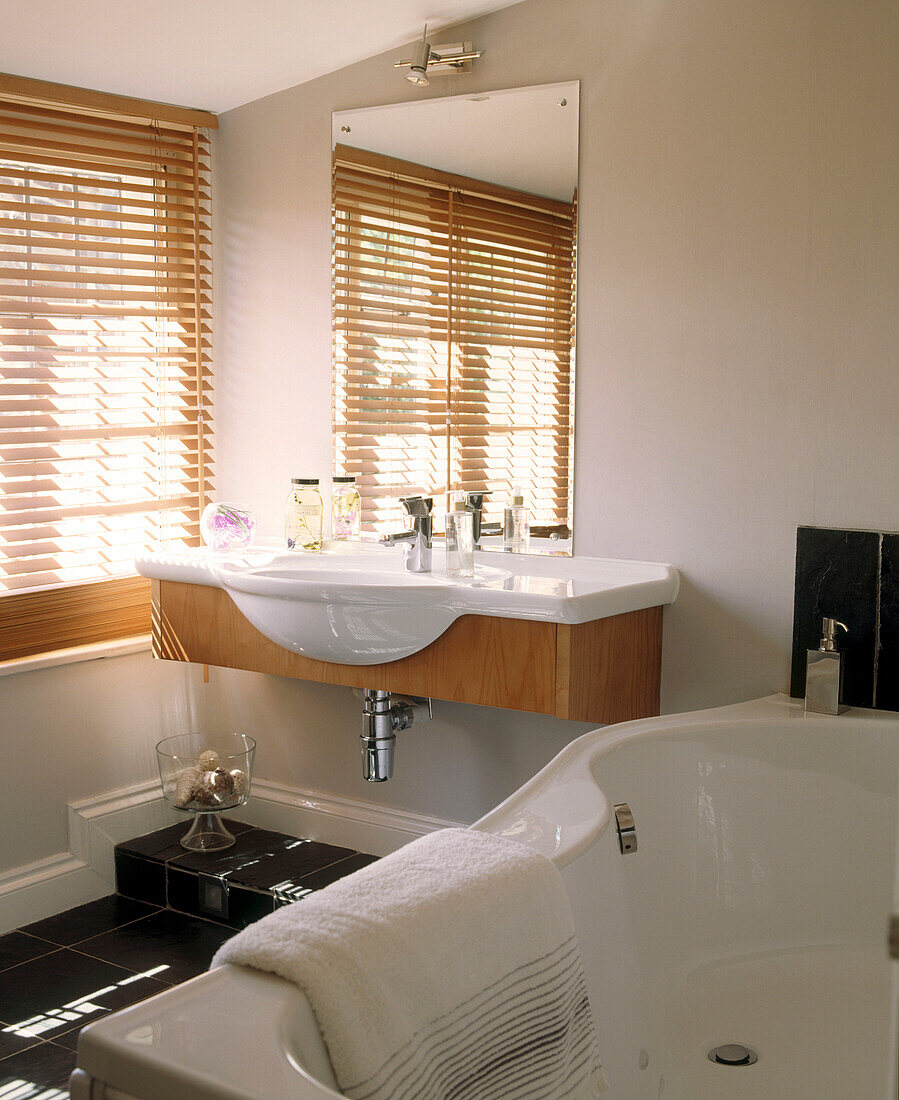 Modernes, in neutralen Farben eingerichtetes Badezimmer mit wandmontiertem Waschbecken in einem Holzschrank, Spiegel, Badewanne und Fenster mit Jalousien