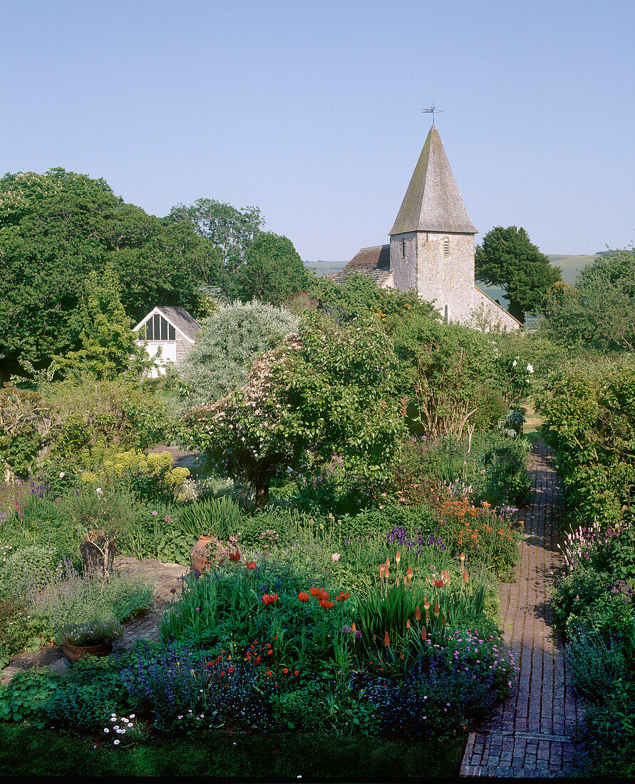 Blick auf einen ländlichen Garten mit Steinkirchturm, Blumenrabatten, Sträuchern und Bäumen