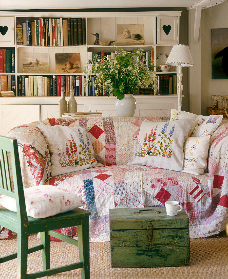 Wohnzimmer im Landhausstil mit Polstersofa mit Patchworkbezug, Holztruhe, gestrichenem Stuhl, Regal, Lampe und Blumenarrangement