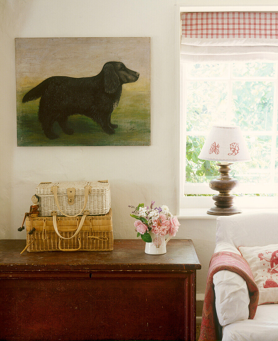 Detail eines Wohnzimmers im Landhausstil, Beistellschrank aus Holz, Karo-Rollo am Fenster gepolstertes Sofa, Weidenkörbe, Gemälde von Hund, Blumenarrangement, Lampe