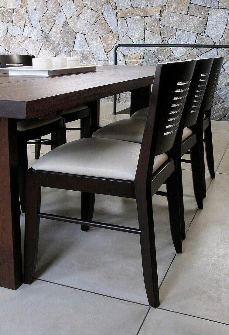 Tisch mit dunklen Holzstühlen auf poliertem Zementboden dahinter Steinwand