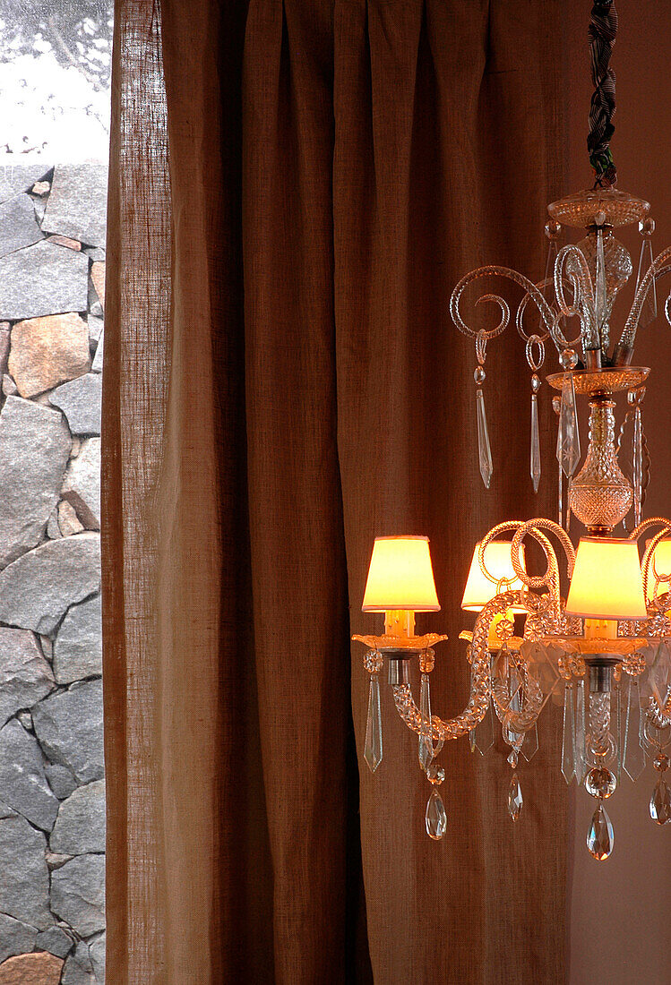 Chandelier hangs against curtains and exposed stone wall