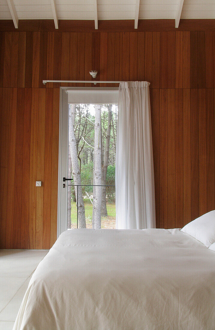 Modernes, mit Zedernholz verkleidetes Schlafzimmer mit Betonboden und Glastür zum Balkon mit Blick auf den Waldgarten