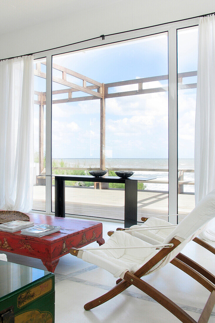Riesige Fenster mit Blick auf das Meer, Leinenvorhänge und ein rot lackierter chinesischer Holztisch mit goldenen Motiven, Sofas und Liegestühle aus weißem Leinen
