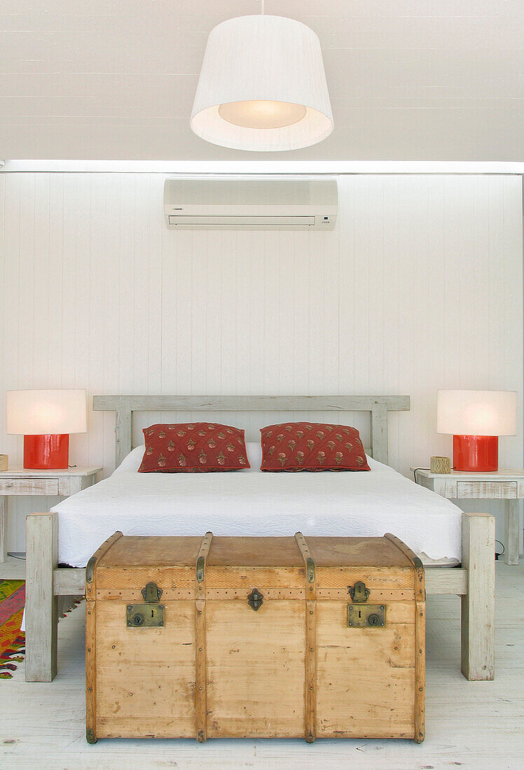 Weiße Pikee-Bettdecke mit roten Kilim-Kissen in einem Schlafzimmer mit Reisekoffer und Hängelampe aus Ripsbändern
