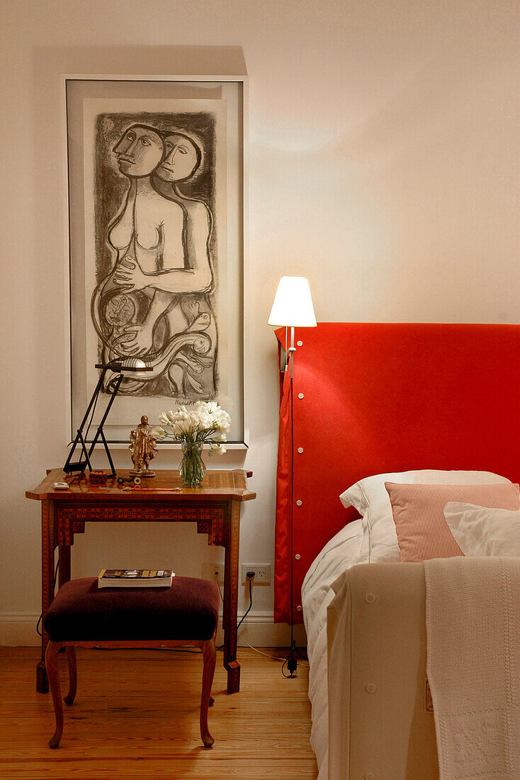 Lit lamp on red headboard in bedroom with modern art above wooden bedside table