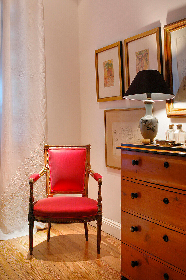 Red upholstered chair with chest of drawers in corner of room with art work