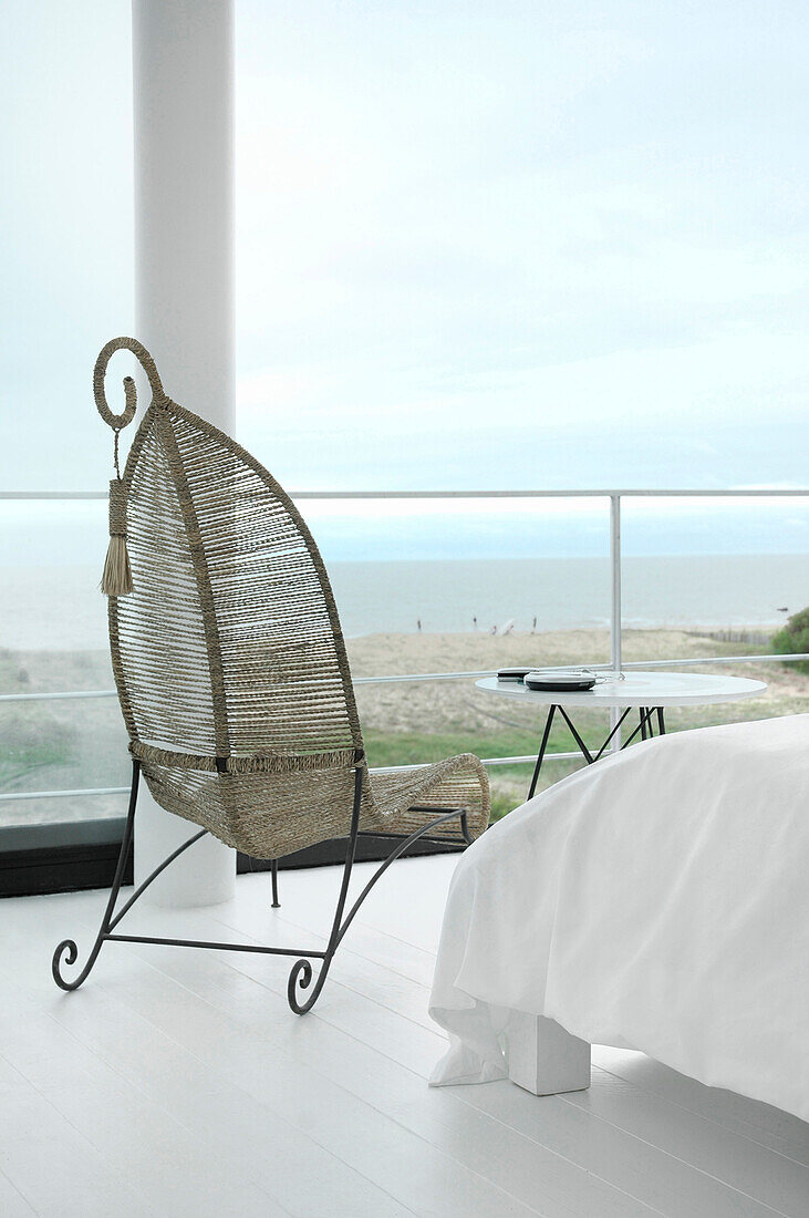 Cane metal framed chair in white bedroom interior with large windows and view to sea