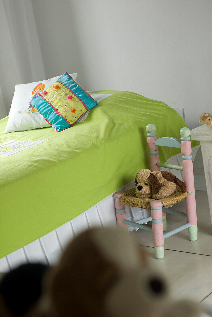 Plüschtier auf bemaltem Stuhl neben einem Kinderbett mit lindgrünem Bezug
