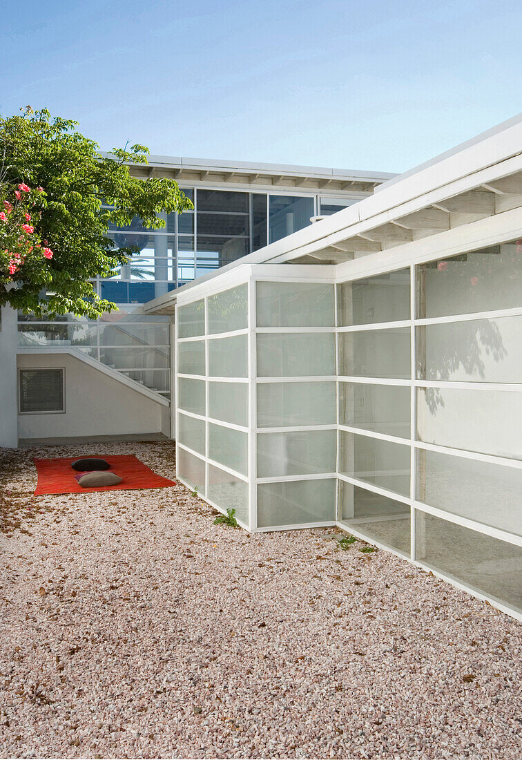 Glass panelled building exterior with gravel courtyard and floor cushions under tree