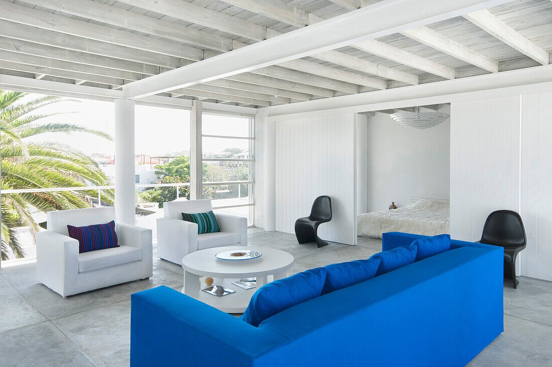 Weißes Wohnzimmer mit großen Fenstern, lebhaftem blauem Sofa und Schiebetüren zum Schlafzimmer