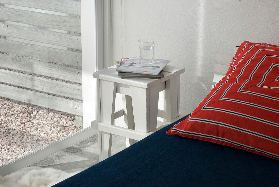 Weißer Nachttisch mit Trinkglas, Zeitschriften und einem roten Kissen