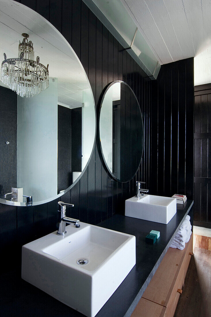 Doppelwaschbecken unter runden Spiegeln in einem schwarz getäfelten Badezimmer mit Kronleuchterleuchte