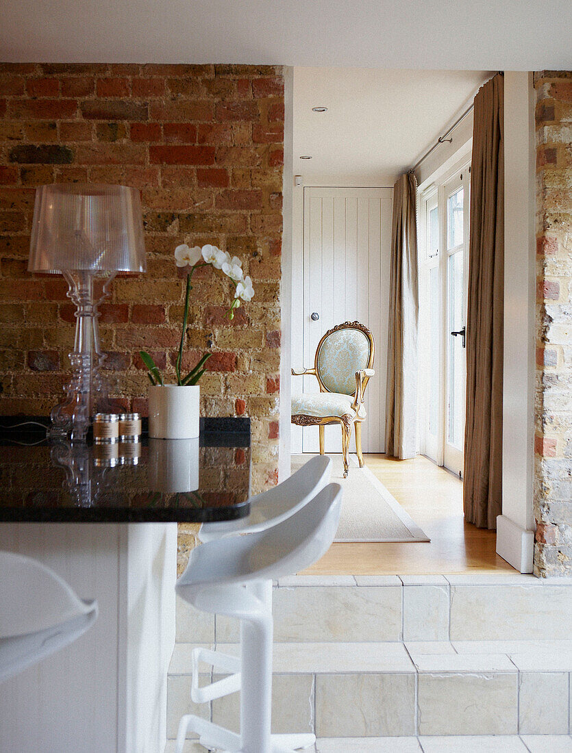 Blick durch die Küche in Richtung Wohnzimmer mit freiliegender Ziegelwand und Retro-Barhockern, weißer Orchidee und Kartel Bourgie Tischlampe mit Louis XV Sessel im Hintergrund