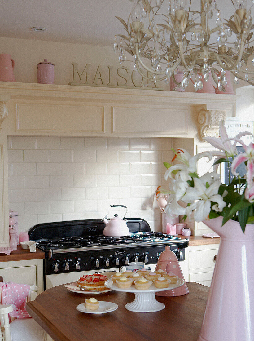 Vase mit Lilien und Törtchen auf Ständer in ländlicher Küche mit rosa Accessoires