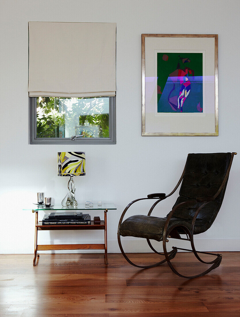 Lederschaukelstuhl mit gläsernem Beistelltisch unter einem Fenster mit moderner Kunst