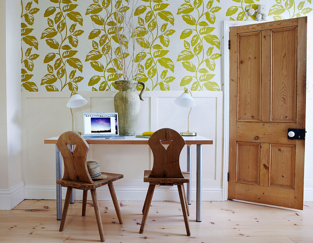 Wooden chairs at study desk with laptop below leaf motif wallpaper