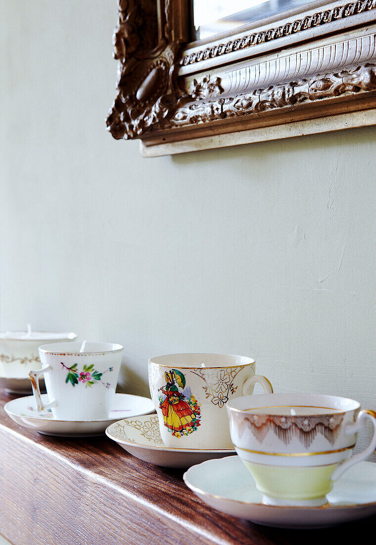 Four teacups with saucers on mantlepiece below carved wooden mirror frame
