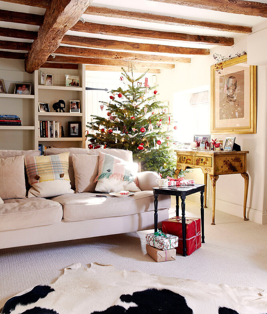 Christmas tree and bookcase with cream sofa in beamed living room of country home