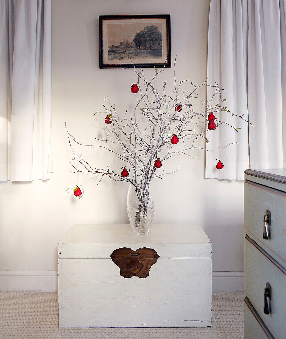Rote Christbaumkugeln hängen an einem Zweigarrangement in einer Glasvase auf einer Reisetruhe