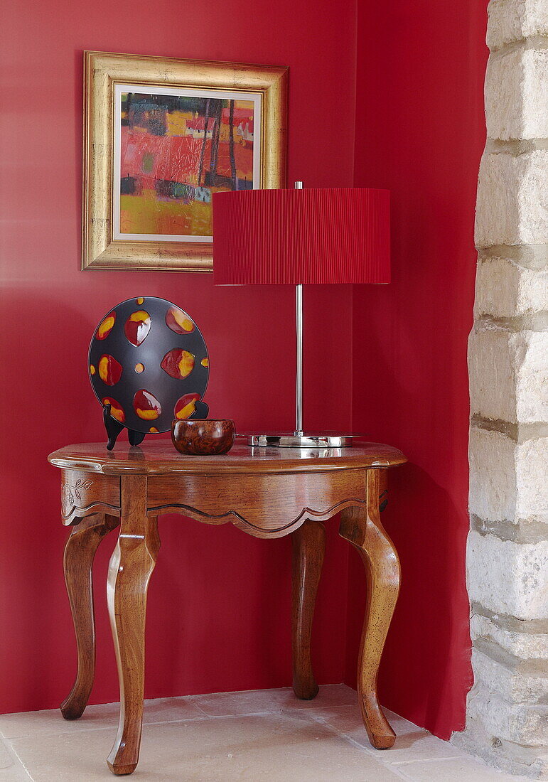 Artwork and lamp on side table with bright red wall in renovated Cotswolds mill house England UK
