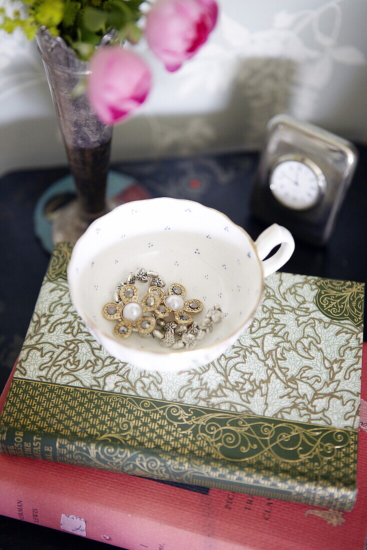Jewellery in teacup with hardbacked books at bedside in Oxfordshire home, England, UK
