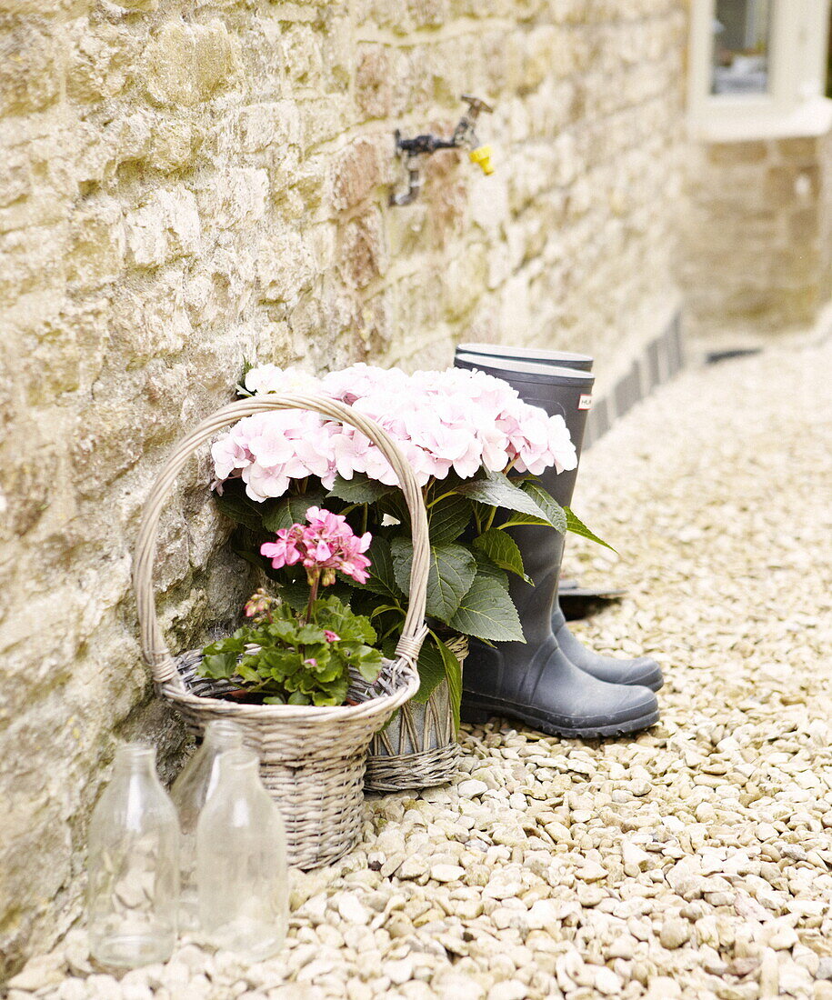 Gummistiefel und Milchflaschen mit blühenden Pflanzen und Wasserhahn an der Steinfassade eines Cottages in Oxfordshire, England, Vereinigtes Königreich