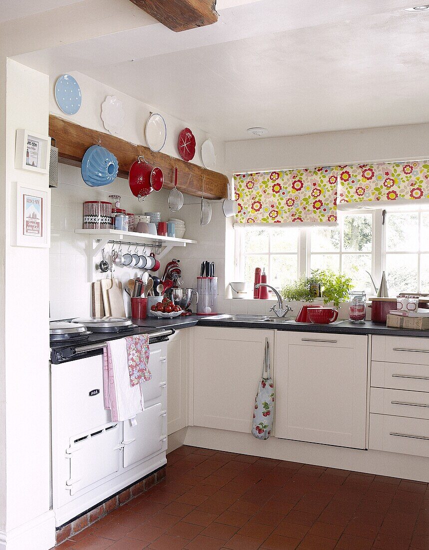Bright red and blue kitchenware in white contemporary cottage kitchen with floral roller blinds in Staffordshire home, England, UK