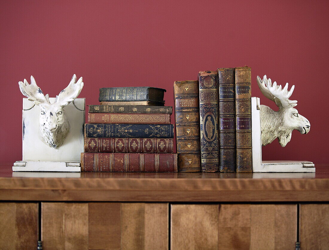 Gebundene Bücher mit Rentierbuchstützen auf Holzschrank vor roter Wand, Oxfordshire, England, UK