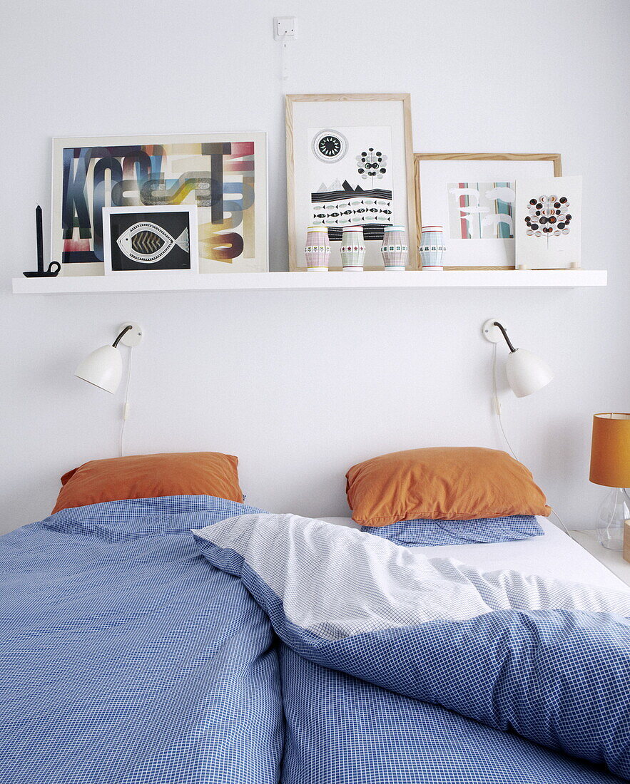 Blau karierte Bettdecke mit orangefarbenen Kissen unter einem Regal mit Kunstwerken im Schlafzimmer eines Hauses in Bussum, Niederlande