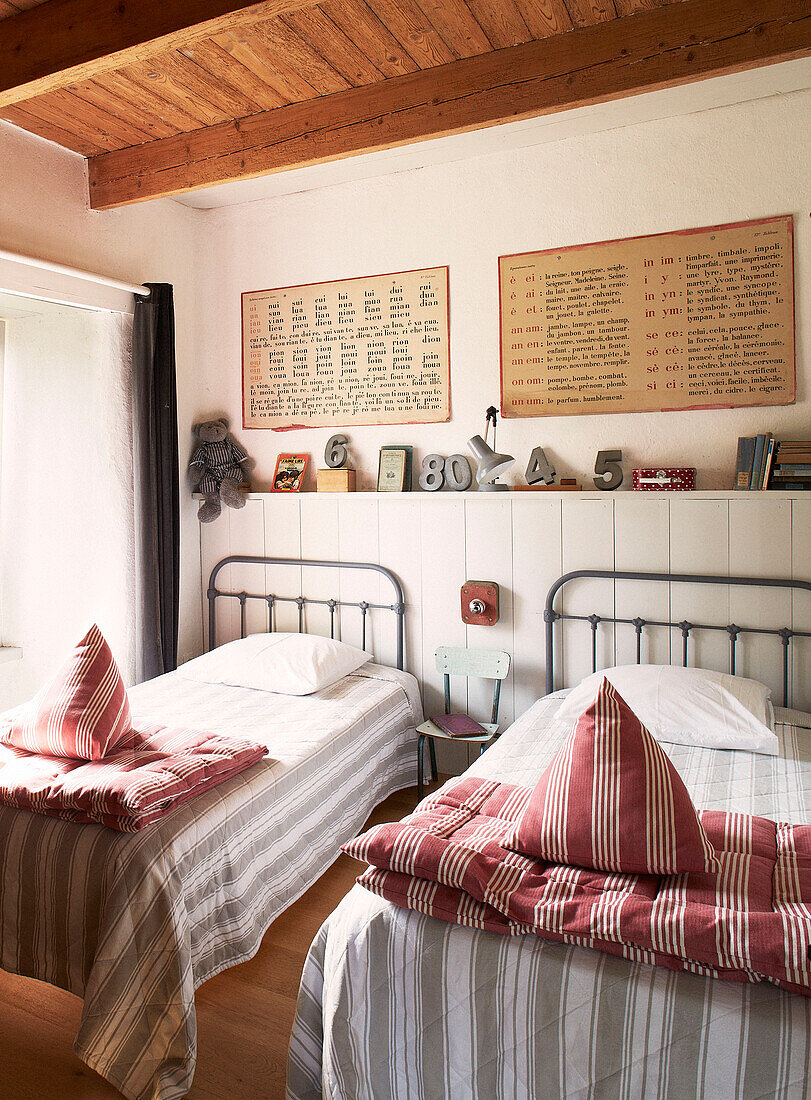 Zwillingsbetten mit gestreifter Bettwäsche unter einem Regal mit Spielzeug und Kunstwerken in einem umgebauten Schulhaus in der Bretagne, Frankreich
