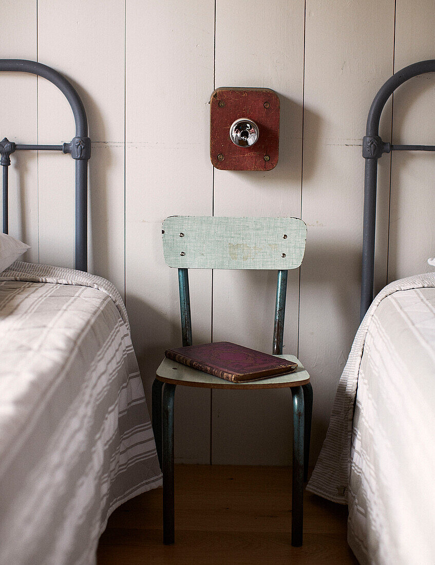 Vintage-Stuhl und Buch zwischen Metall-Kopfteilen im Zweibettzimmer eines umgebauten Schulhauses in der Bretagne Frankreich