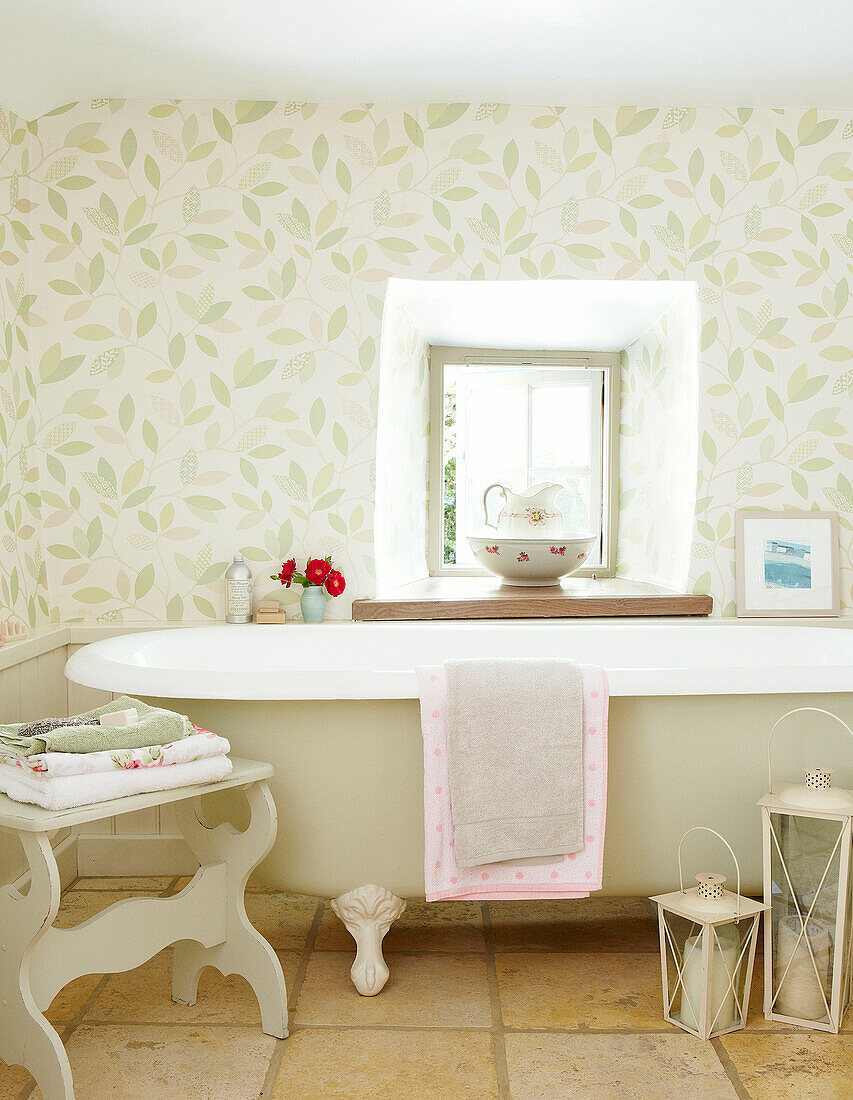 Freestanding bath below window with leaf patterned wallpaper in Devonshire cottage UK