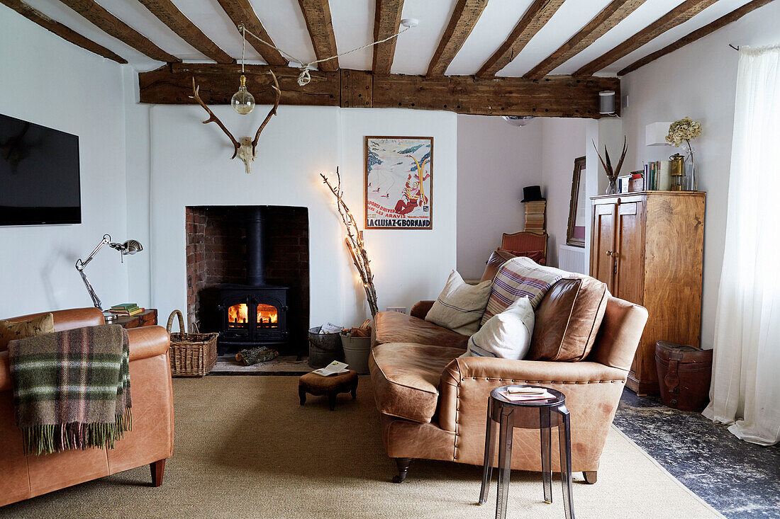Brown leather sofa and chair with lit woodburner in beamed Warwickshire farmhouse, England, UK