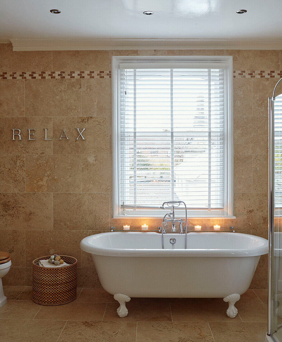 Freestanding candlelit rolltop bath at window with Venetian blinds and single word ' RELAX' in, UK home