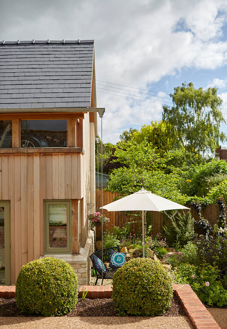 Abgeschnittene Buchsbaumkugeln und Sonnenschirm im Garten eines holzverkleideten Neubaus in den Cotswolds, UK