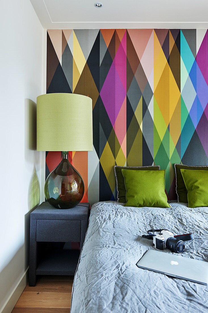 Doppelbett und Nachttisch mit Lampe vor Wand mit buntem, grafischem Muster