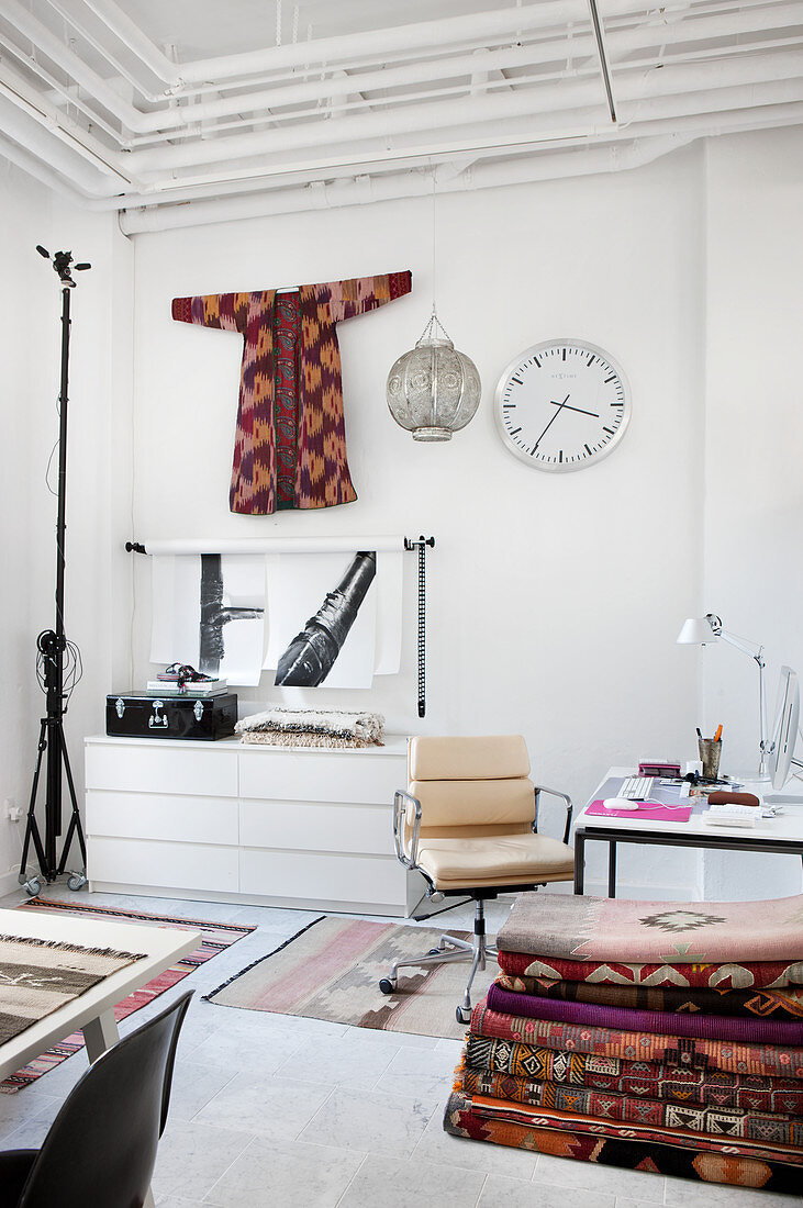 Weiß gestrichenes Arbeitszimmer mit Klassikerstuhl, gestapelten Kelimteppichen und Kimono als Wanddekoration