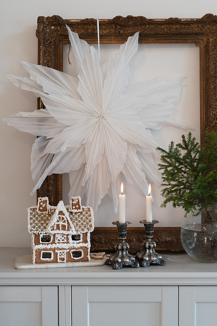 Gingerbread house and candles below paper star in gilt picture frame