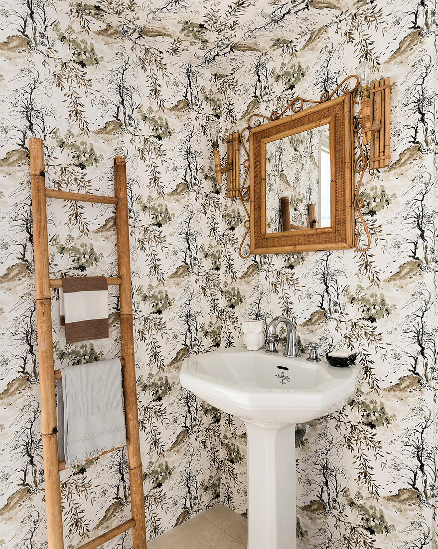 Bamboo mirror above pedestal sink and ladder in bathroom with patterned wallpaper