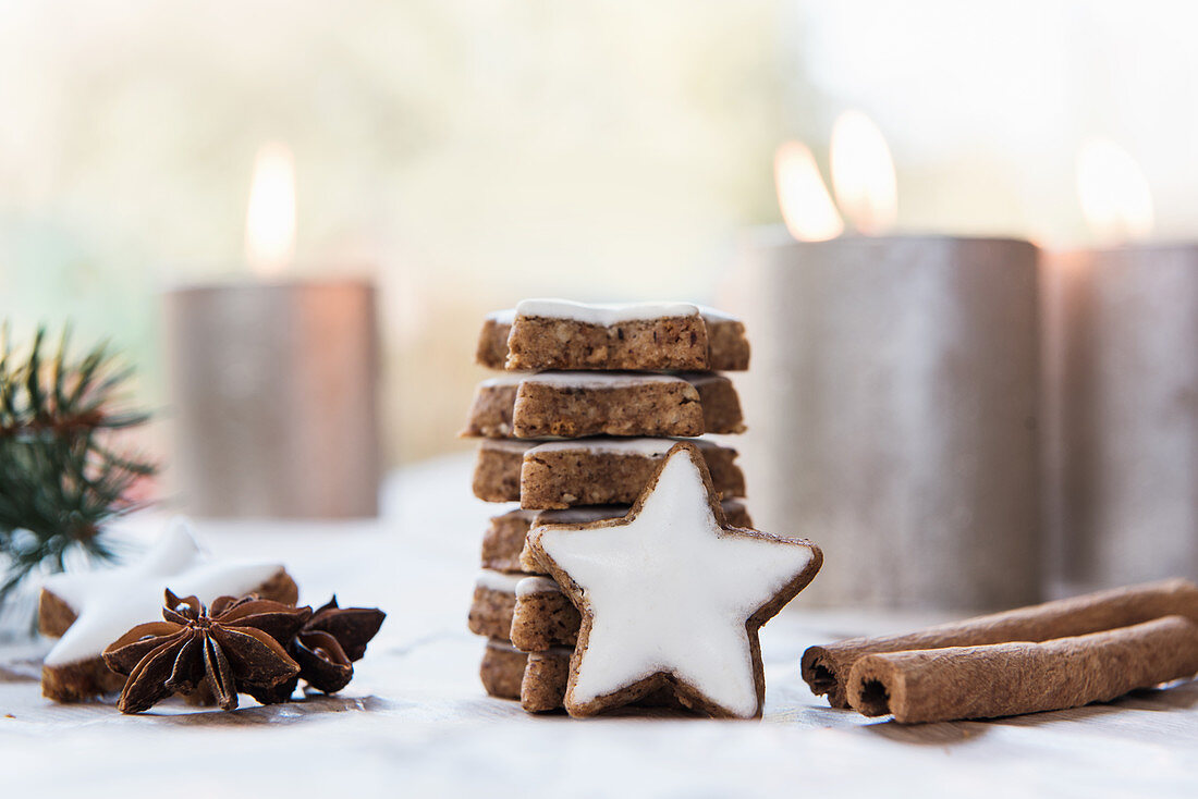 Stack of cinnamon star biscuits, cinnamon sticks, star anise and lit candles in background
