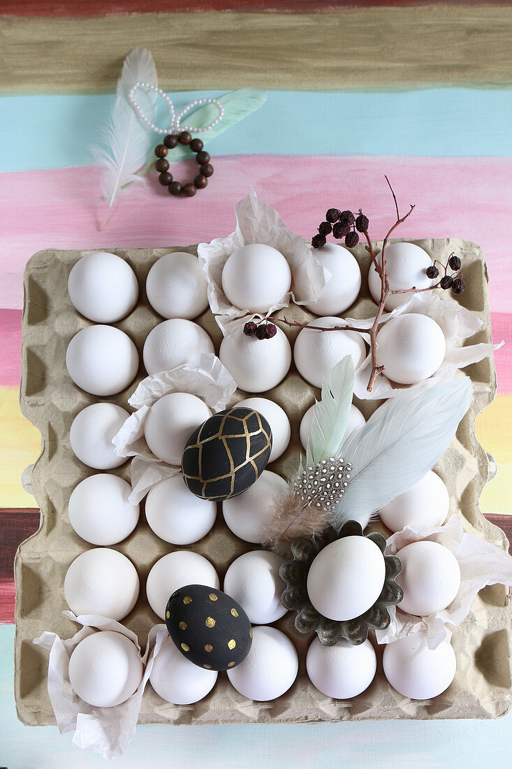 Bemalte Eier, Federn und Zweig auf einer Eierpalette mit weißen Eiern