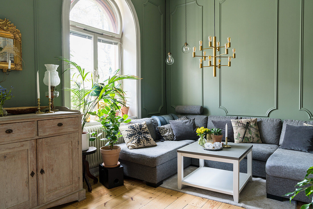 Anrichte, graue Polstergarnitur, Zimmerpflanzen vor Rundbogenfenster im Wohnzimmer mit grünen Wänden