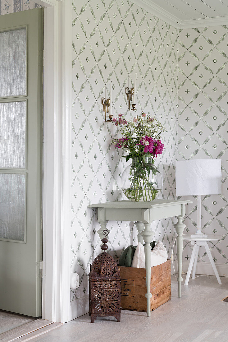 Konsolentisch mit Blumenstrauß und Tischlampe auf Hocker vor tapezierter Wand